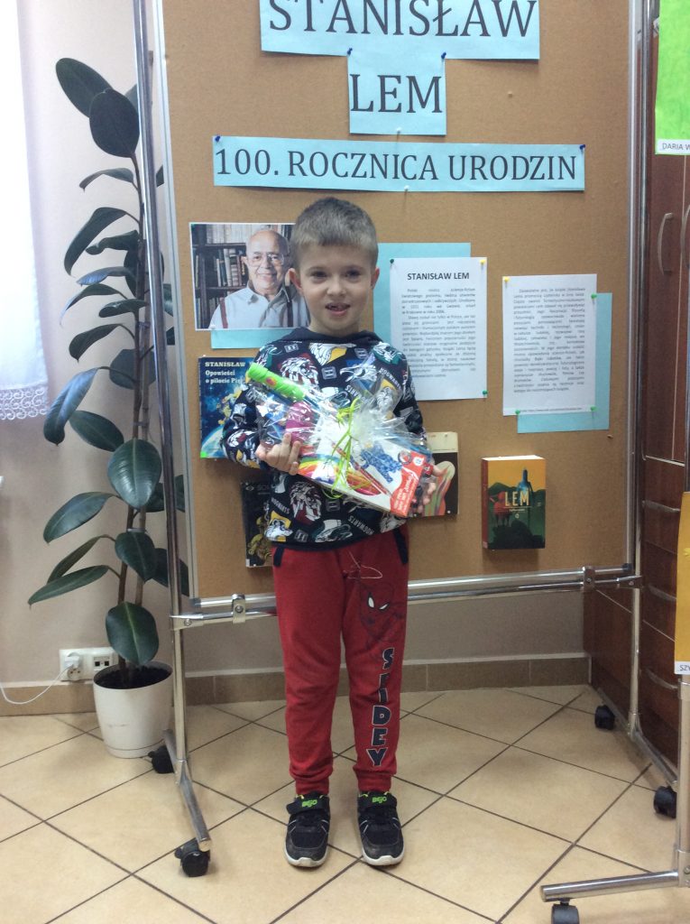 Uśmiechnięty chłopiec stoi i trzyma w rękach pakunek zawinięty w celafon. Za nim tablica z napisami. Po lewej wysoka roślina doniczkowa.