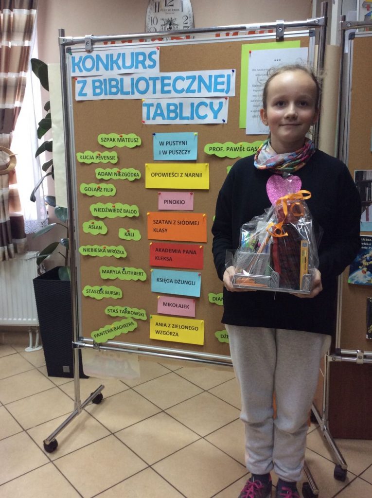 Na pierwszym planie dziewczynka trzymająca zapakowaną nagrodę. W tle tablica z napisem "konkurs z bibliotecznej tablicy".