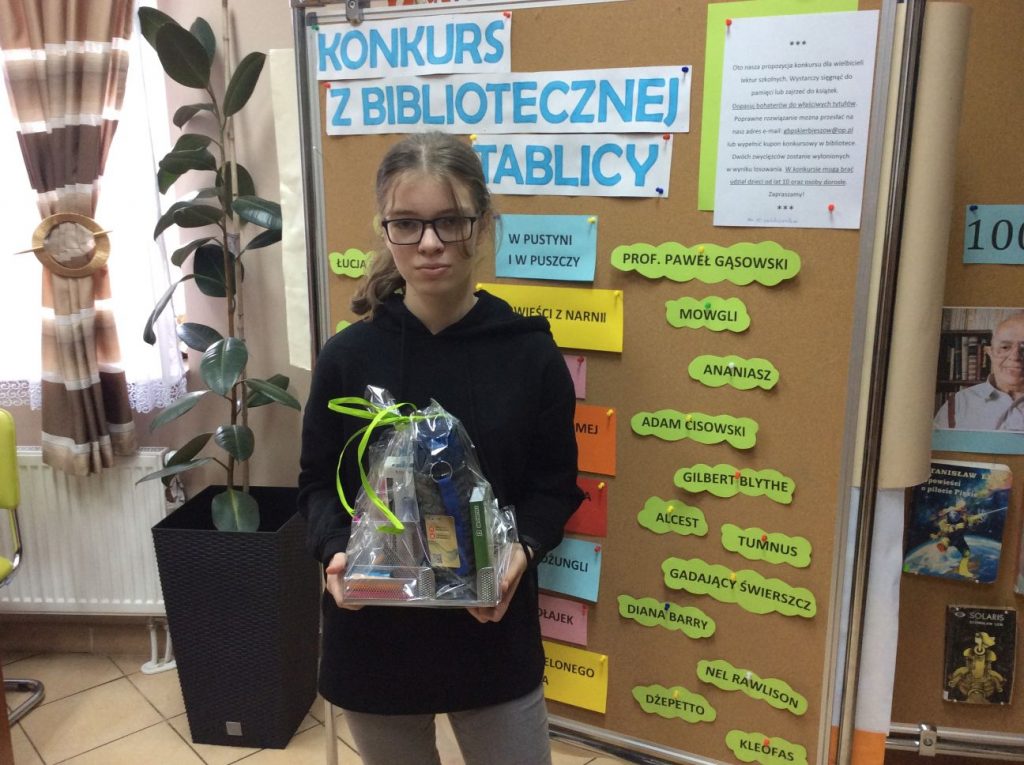 Na pierwszym planie dziewczyna trzymająca zapakowaną nagrodę. W tle tablica z napisem "konkurs z bibliotecznej tablicy".
