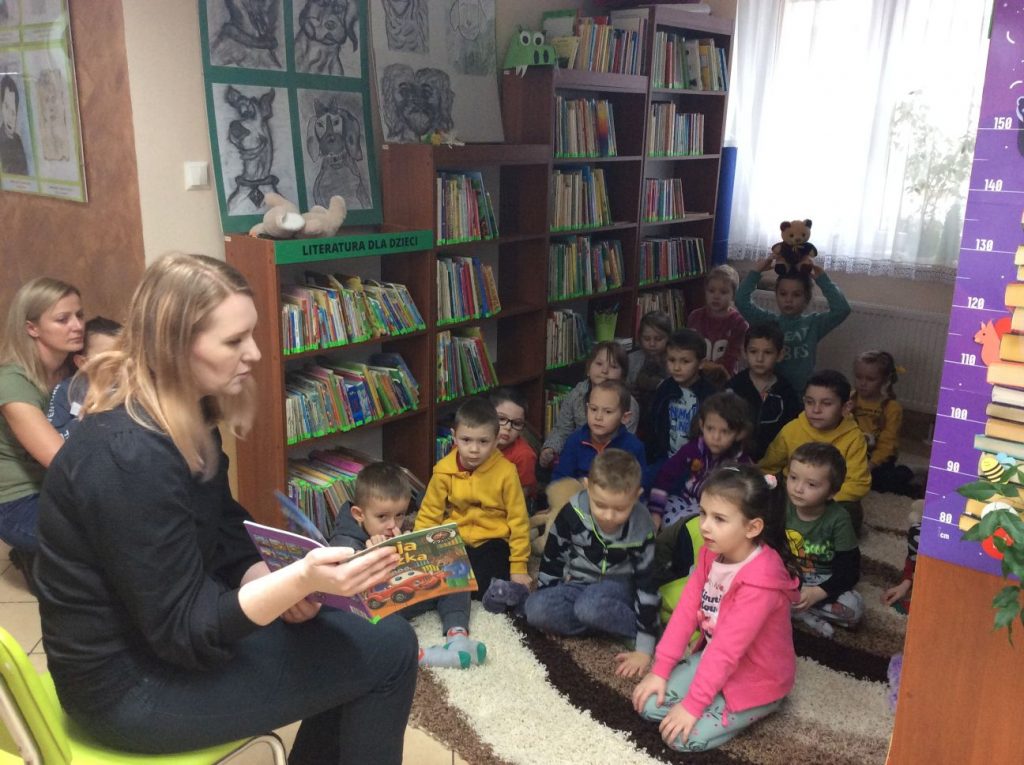 Bibliotekarka czyta dzieciom książeczkę. Dzieci siedząc na dywanie słuchają czytanego opowiadania.