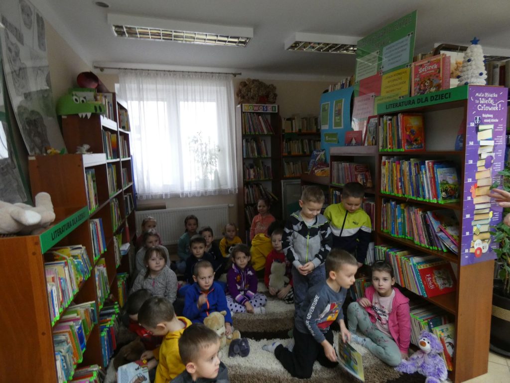 Pomiędzy regałami wypełnionymi książkami na dywanie siedzą dzieci w wieku przedszkolnym.