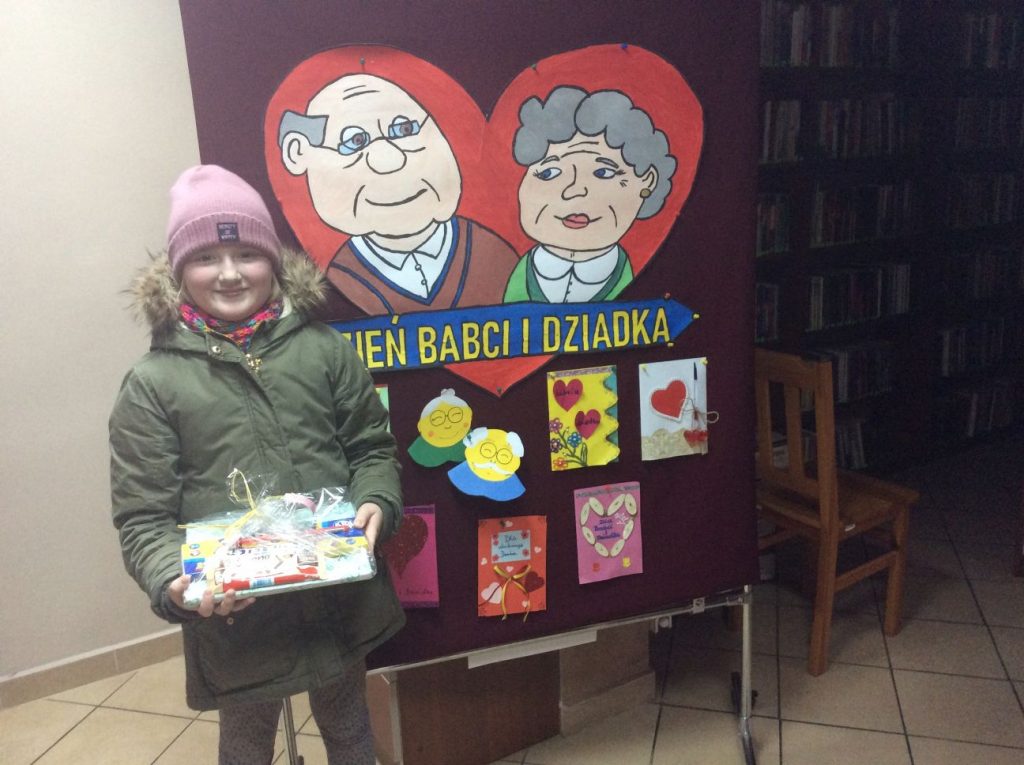 Na tle gazetki okolicznościowej z okazji Dnia Babci i Dziadka stoi uśmiechnięta dziewczynka z nagrodą.