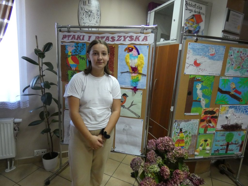 Dziewczynka stoi na tle tablicy  przedstawiającej prace plastyczne - kolorowe ptaki. Po lewej stronie na podłodze ustawiono bukiet z hortensji.