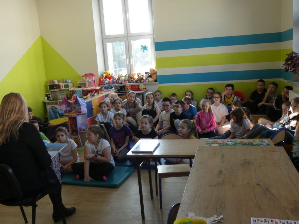 Duża grupa dzieci siedzi na materacach w klasie. Przed nimi bibliotekarka czytająca książkę.
