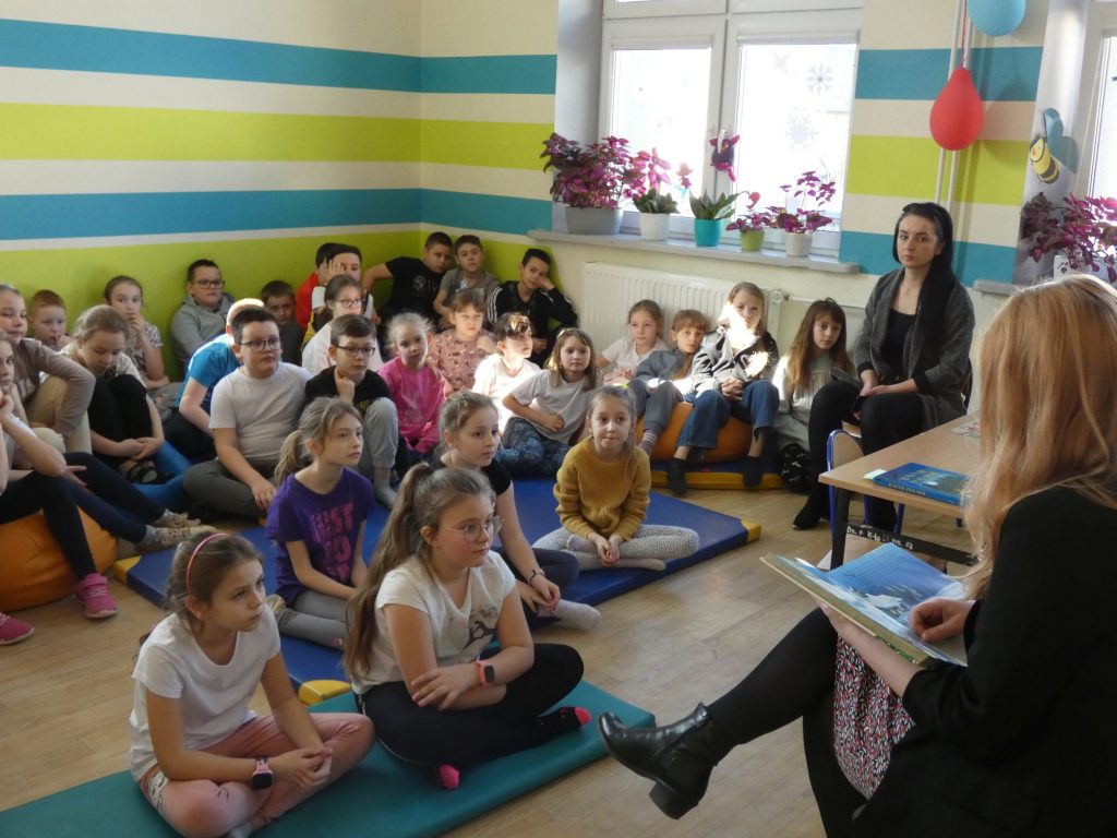 Duża grupa dzieci siedzi na materacach w klasie. Przed nimi bibliotekarka czytająca książkę. Obok dzieci po prawej  - siedzi nauczycielka.