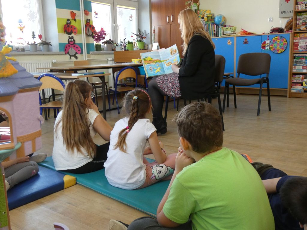 Na tle klasy szkolnej bibliotekarka siedząc na krześle czytająca kolorową książkę dla dzieci. Przed nią czworo dzieci siedzących na materacach.