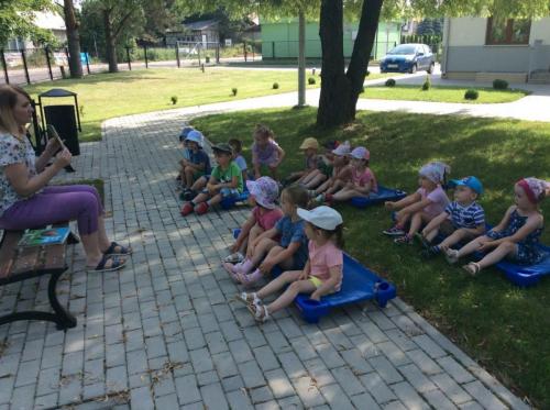 Letnia sceneria, zielony trawnik. Kilkunastoosobowa grupa dzieci w wieku przedszkolnym siedzi na leżaczkach i  słucha opowiadań czytanych przez bibliotekarkę.