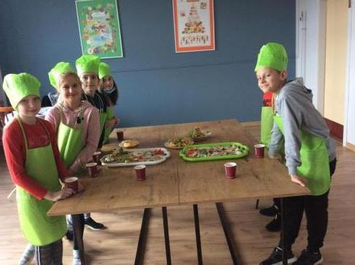 Szóstka dzieci w zielonych fartuszkach i czapkach kucharskich zgromadzona przy stole zastawionym przygotowanymi przekąskami.