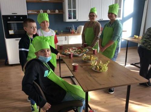 Piątka dzieci w zielonych fartuszkach i czapkach kucharskich zgromadzona przy stole zastawionym przygotowanymi przekąskami.